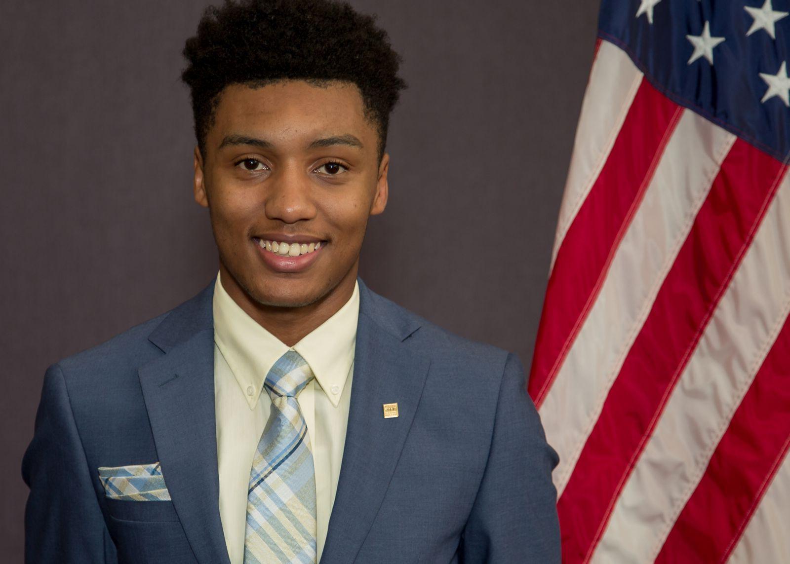 非裔美国大学生Rah'Mere Williams, 他身穿深蓝色西装，配上白色领衬衫，系着浅蓝色条纹领带，站在一面灰色背景的美国国旗的前面和右边.